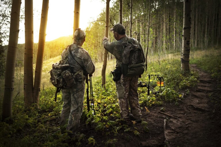 Two bow hunters in full camouflage walking through hunting land for sale toward their tree stand, surrounded by tall trees and natural vegetation.