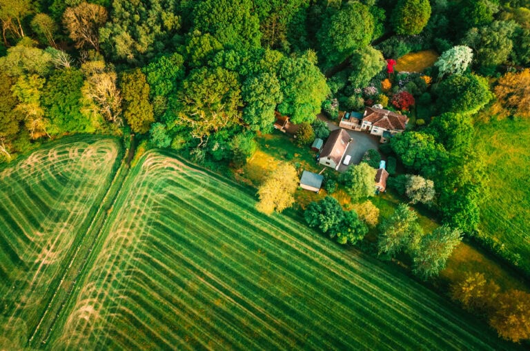 Aerial view of a rural home surrounded by wooded acreage and backing up to open farm fields, illustrating the unique characteristics and land-use considerations involved in buying land versus residential property.